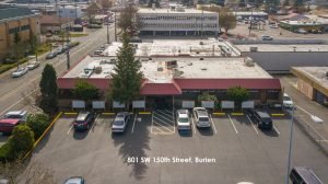 An aerial view of a city block with a parking lot and cars outside a flat-roofed, one-story building with red awnings.