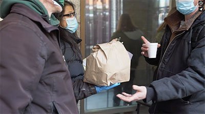 A person in a face mask hands a filled brown paper bag to another person in a face mask.