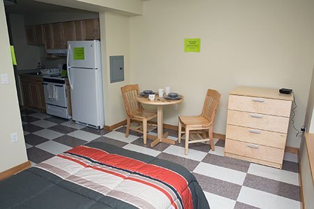 A view from the right of the end of a bed covered with a gray-and-red-striped comforter, to a light-colored bistro table, chairs and chest of drawers, and part of a kitchenette, with refrigerator and stove.