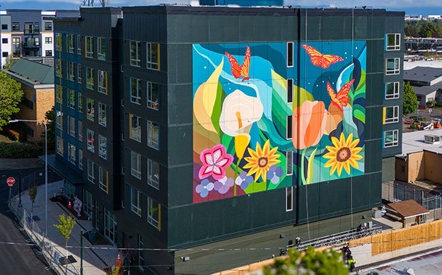 The main subject is a building painted black and covered all along one wall with a vibrantly colored four-story high mural of flowers and butterflies. Artwork by Angelina Villalobos; photo courtesy of barry johnson, overall creative.